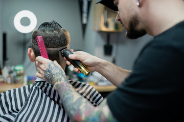 A barber man with a tattoo on his arm makes a haircut to a brunette guy in a barbershop, cuts his...