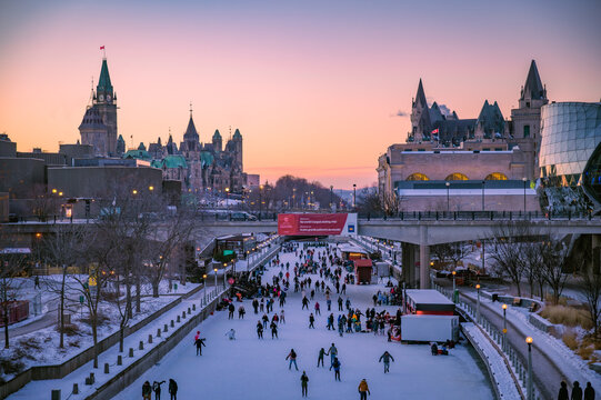Downtown Ottawa during winter with skaters on the Rideau Canal Skateway, world's largest skating rink, with Parliament Hill and Fairmont Chateau Laurier, sunset dusk sky, Ontario, Canada (January 2022