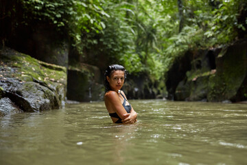 A young slender woman in a swimsuit poses by a mountain river in the jungle on the popular island of Bali.
