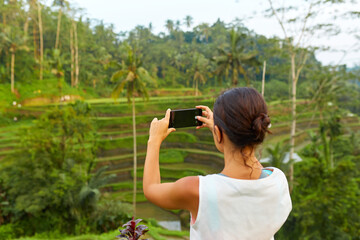 A young slender woman stands with phone on rice terraces on the popular tourist island of Bali.