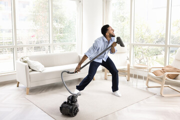 Excited funny young African homeowner man singing at hoover tube like at microphone, cleaning floor...