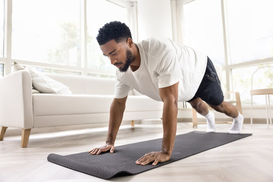 Focused athletic strong African man doing pushups, morning exercises at home, caring for power, strength, wellness, sport, training body, keeping static plank pose on yoga mat - Powered by Adobe