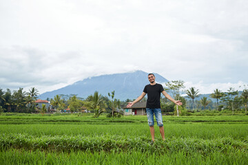 Naklejka na ściany i meble A young man poses in rice fields against the backdrop of Mount Agung volcano on the popular tourist island of Bali.