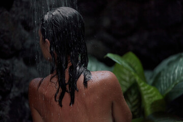 A naked woman takes a shower in an outdoor shower with tropical plants. A shampoo advertisement in dark colors.