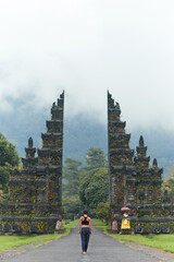 A woman poses at the Balinese traditional gate against the background of mountains with clouds on...