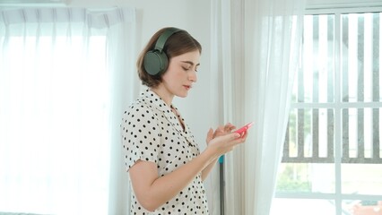Caucasian mother put on headphone while choosing song by using mobile phone before doing housework. Headset placed on table while was putting on happy housekeeper to listen relaxing song. Pedagogy.