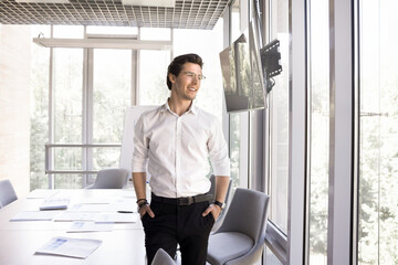 Pensive smiling millennial 35s businessman standing alone in modern conference room think on...