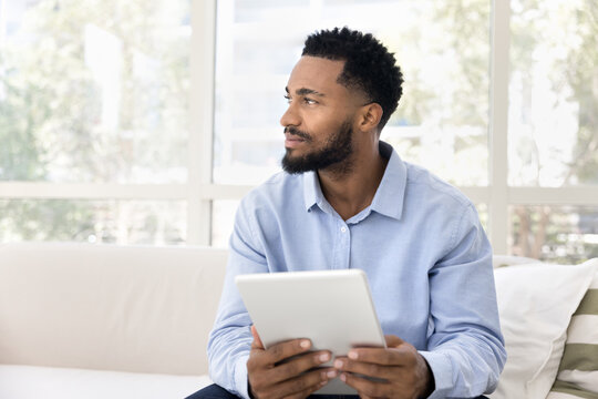 Positive thoughtful young African American man using digital tablet computer at home, holding gadget, looking away, thinking on Internet communication, freelance work project