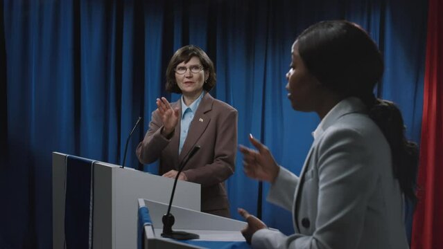 Medium close-up side shot of agitated young African American female politician giving angry speech at election debate, blaming economic crisis on opponents party, who is trying to defend herself