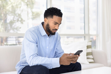 Focused young handsome Black man typing message on cellphone phone, sitting on sofa at home, using...