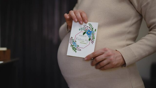 Close-up. Slow motion. Happy Mother's Day. Profile of an unrecognizable pregnant woman, holding a Mother's Day card with one hand and rubbing her belly with the other hand, while standing indoors