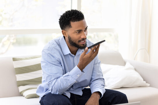 Serious young Black business man holding smartphone at mouth, dictating voice command at speaker, speaking audio message at mobile phone, activating virtual assistance app - Powered by Adobe