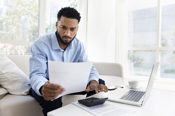 Fototapeta premium Serious young African American man calculating expenses, income at home, typing on calculator, using laptop, reading legal financial document, doing accounting paperwork