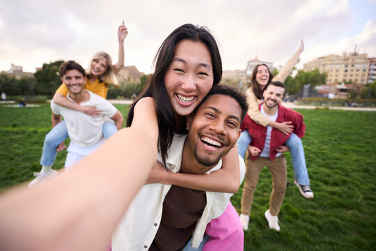 Multirracial group of young smiling friends taking selfie picture in piggyback in the park hugging by couples. Happy millennial people smiling together looking at camera outside. University students