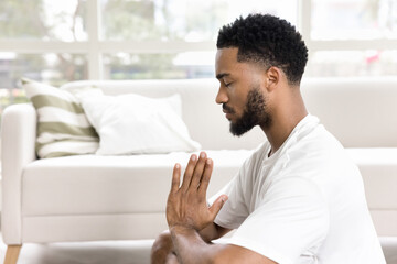 Focused calm African American guy meditating at home, practicing pranayama, breath work, keeping Namaste greeting hands gesture, sitting on floor with closed eyes. Side view - obrazy, fototapety, plakaty