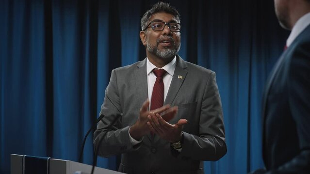 Medium footage of cheerful, expressive middle-aged Indian male political candidate in suit, tie taking off glasses and patronizing unrecognizable adversary, while attending election debate