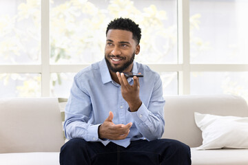 Positive handsome young African business man talking on speaker on cellphone, recording voice message, looking away, thinking, activating modern smart home application