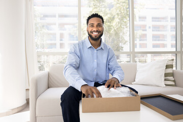 Happy handsome young Black influencer guy unwrapping parcel, opening cardboard package, reviewing...