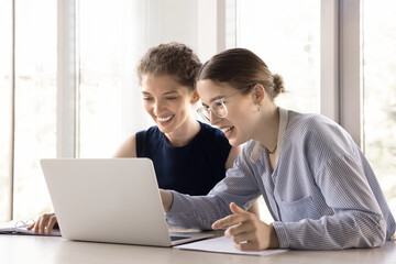 Two young happy office employees, women colleagues, teammates sit at desk with laptop, talking,...