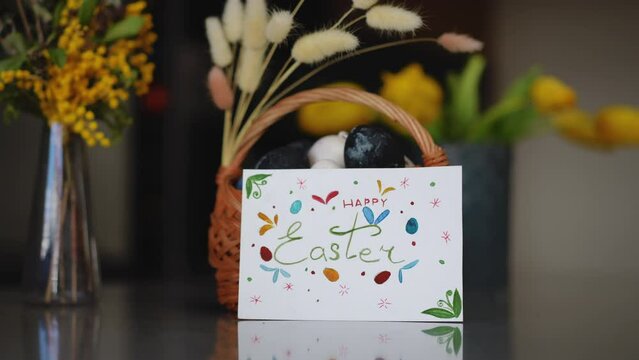 Close-up. Happy Easter holiday. On the indoor table is a glass vase with spring flowers and a decorated basket with Easter eggs. A woman's hand places a handmade Easter card near a basket