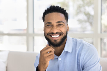 Happy joyful handsome young African guy looking at camera with toothy smile, touching black stubble, sitting on couch, talking on video call. Successful business professional head shot portrait