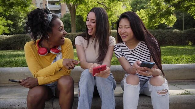 Three joyful multiracial women using phone sitting outdoor in summer. Excited girl shows something funny on mobile to friends. Young female laughing in surprise looking and pointing at screen device