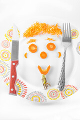 A smiling face made from slices of a carrot, grated carrot hair and a beard of green leaves on a white plate with colorful circle patterns. A kitchen knife and a fork on each side.