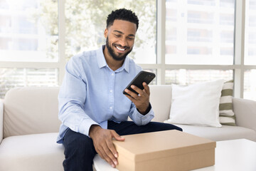 Happy young African American client man typing on mobile phone over logistic cardboard box, receiving parcel from delivery transportation service, using smartphone, smiling, laughing