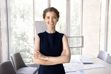 Fototapeta na wymiar Beautiful young businesswoman standing alone with arms-crossed in modern boardroom. Elegant millennial businesslady, coach or business trainer, talented designer smile look at camera posing in office