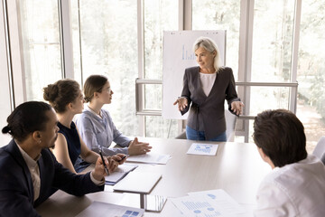 Multiethnic team listen female business trainer gives presentation for businesspeople gathered in modern conference room. Mature businesslady lead seminar or educational workshop for company employees