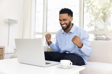 Cheerful excited African American freelance employee man celebrating job success, sitting on couch at coffee table, making winner yes fists gesture at laptop computer, laughing