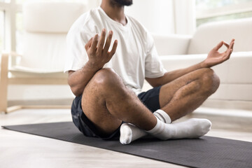 Cropped shot of young African man meditating after yoga practice, sitting on mat on floor, keeping zen hands gesture, doing breath work, caring for concentration, mindfulness, calmness, harmony