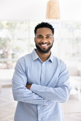Happy handsome young African American entrepreneur man in office shirt standing indoors with crossed arms, looking at camera with toothy smile, laughing. Business professional vertical portrait