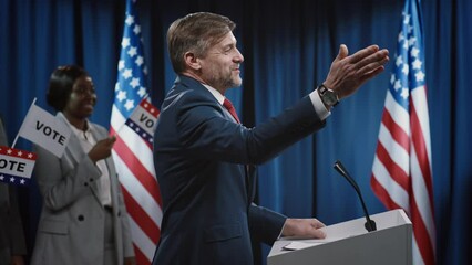 Medium side shot of middle-aged Caucasian male US president or senator walking onto stage towards podium, greeting audience and delivering energetic keynote address while campaigning for reelection