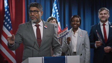 Medium shot of charismatic Indian American male candidate for Senate or US President delivering political speech on stage at conference or convention, interacting with public, photo cameras flashing