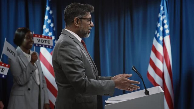 Medium side footage of middle-aged biracial Indian American male candidate for president or governor from Republican party walking up to podium and greeting public in hall, photo cameras flashing