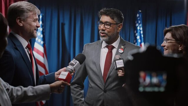 Medium shot of multiethnic journalists with microphones and camera interviewing charismatic Indian American male presidential candidate, and asking questions on electoral programme, migration policy