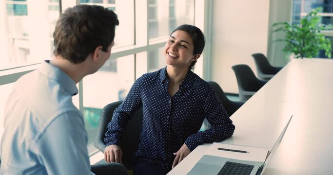 Glad Indian businesslady shake hand client after successful meeting in office, sit at desk with laptop, make agreement, closing commercial deal, enjoy teamwork express gratitude with handshake gesture
