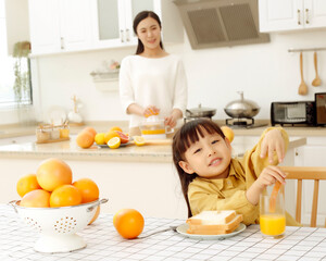 Asian woman making orange juice in kitchen, girl drinking juice and eating bread