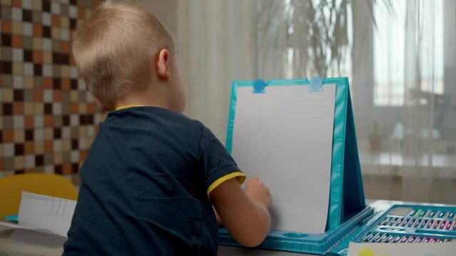 A little boy draws with a pencil on a piece of paper at home. Back view of a blond, focused child learning to draw simple shapes while studying at home. Early development and homeschooling concept