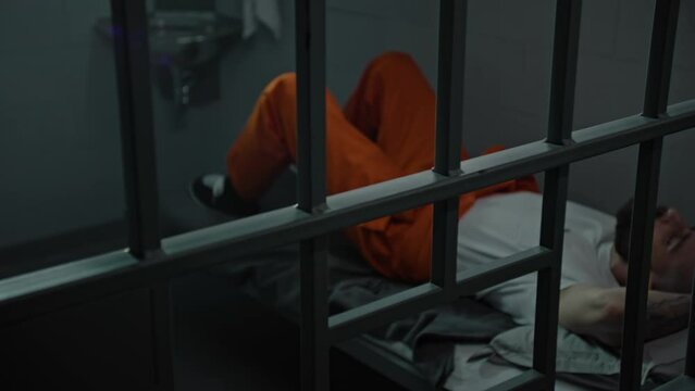 Inmate in orange uniform lies on prison cell bed. Prisoner serves imprisonment term for crime. Criminal in detention center, correctional facility. Justice system. View through metal bars. Dolly shot.