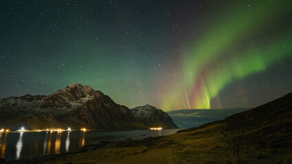 Aurora borealis over the clouds above the sea and fishing villages by the mountains. Lofoten Islands, Northern Norway.