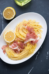 Spaghetti served with prosciutto and lemon zest on a white plate, vertical shot on a black marble background, high angle view