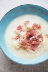 Middle close-up of potato soup served with torn prosciutto in a turquoise bowl, vertical shot, selective focus
