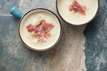 Bowls of potato soup with prosciutto on a beige and grey granite background, horizontal shot with space, above view