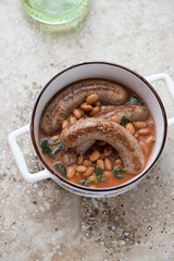 White serving pan with roasted sausages and borlotti beans, vertical shot on a beige granite background, high angle view