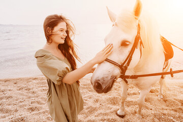 A woman in a dress stands next to a white horse on a beach, with the blue sky and sea in the...