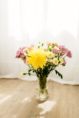 A bouquet of yellow and pink asters and gerbers in a glass vase against the backdrop of a window with rays of sun. Flowers in a vase in the interior