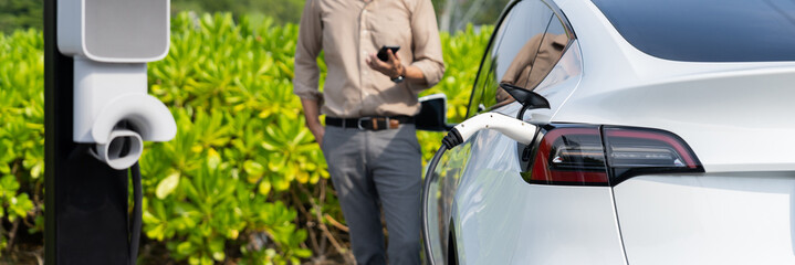 Young man use smartphone to pay for electricity at public EV car charging station green city park....