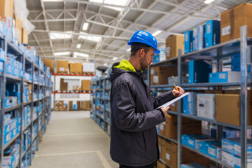 Young caucasian man in helmet, worker in warehouse checking carton boxes goods. Stock of Parcels with Products, transportation concept